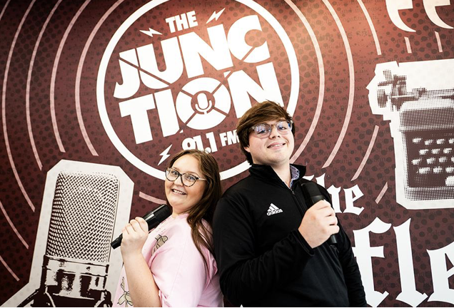 Two MSU students smile and hold microphones in front of a wall in the Junction radio station