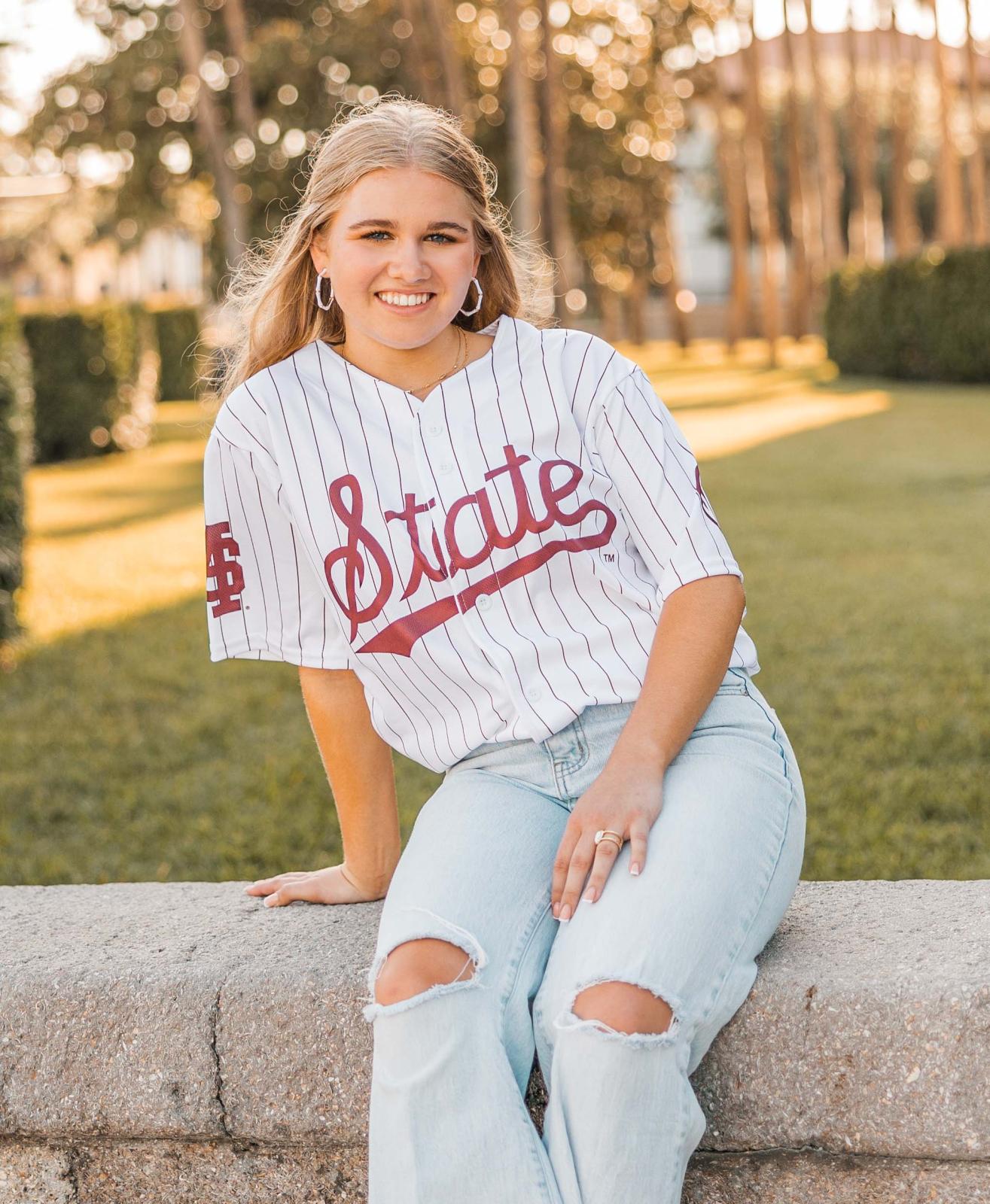 River Neri smiles at the camera. She is sitting on a stone wall wearing jeans and a State jersey.
